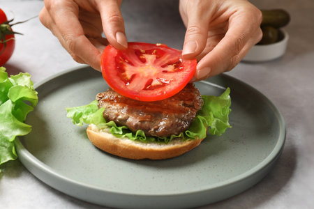 Woman making tasty hamburger with fried patty, vegetables and bun at table, closeupの写真素材