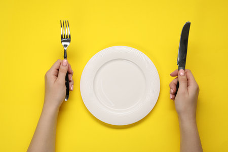 Woman holding fork and knife near empty plate at yellow table, top viewの写真素材