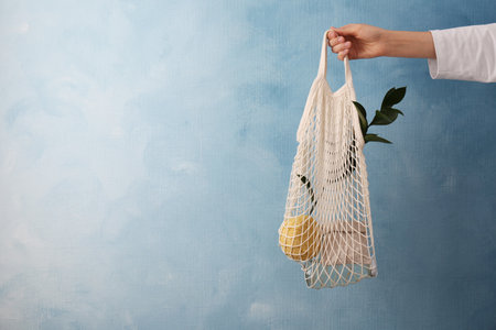 Woman holding net bag with different items against light blue wall, closeup and space for text....