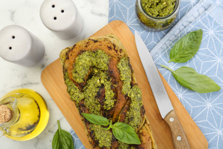 Freshly baked pesto bread with basil and knife on white marble table, flat layの写真素材