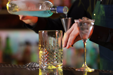 Cocktail making. Bartender pouring alcohol from bottle into jigger at counter in bar, closeupの写真素材