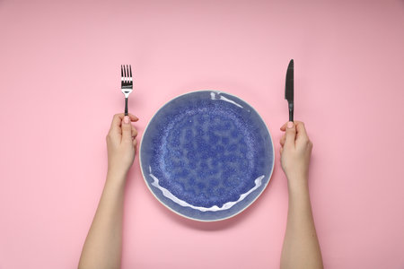 Woman holding fork and knife near empty plate at pink table, top viewの写真素材