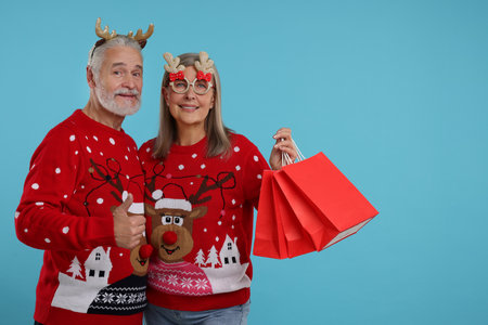 Senior couple in Christmas sweaters, reindeer headband and party glasses with shopping bags on light blue background. Space for textの写真素材