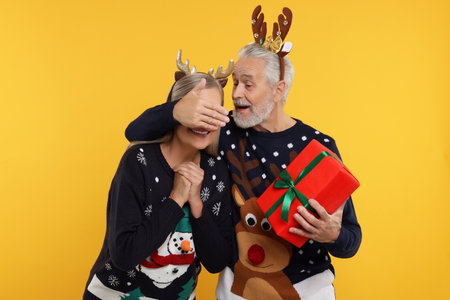Couple in Christmas sweaters. Senior man surprising his woman with gift on orange backgroundの写真素材