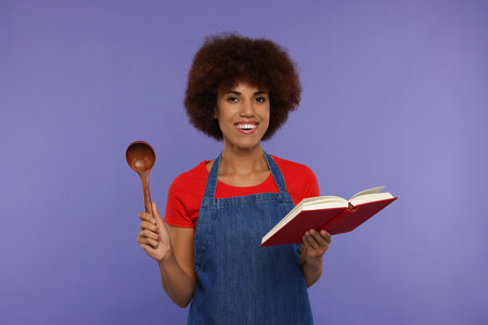 Happy young woman in apron holding spoon and recipe book on purple backgroundの写真素材
