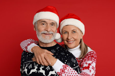 Senior couple in Christmas sweaters and Santa hats on red backgroundの写真素材