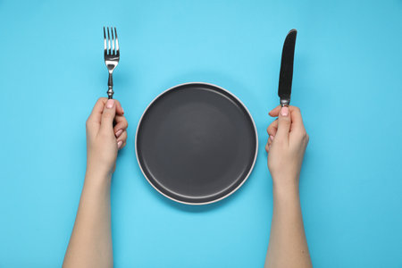 Woman holding fork and knife near empty plate at light blue table, top viewの写真素材