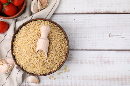 Bowl of raw bulgur with scoop, tomatoes and garlic on white wooden table, flat lay. Space for textの写真素材