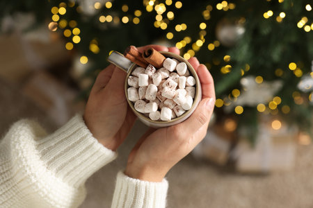 Woman holding cup of delicious Christmas cocoa with marshmallows and cinnamon sticks against blurred lights, top viewの写真素材