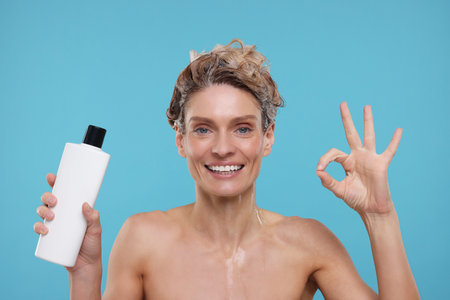 Washing hair. Portrait of happy woman with bottle showing ok gesture on light blue backgroundの写真素材