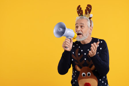 Senior man in Christmas sweater and reindeer headband shouting in megaphone on orange background. Space for textの写真素材