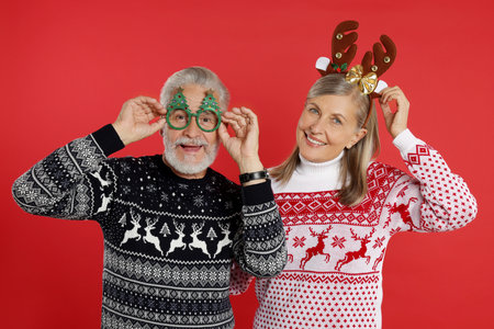 Happy senior couple in Christmas sweaters, reindeer headband and funny glasses on red backgroundの写真素材
