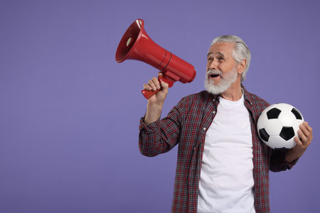 Emotional senior sports fan with soccer ball using megaphone on purple background, space for textの写真素材