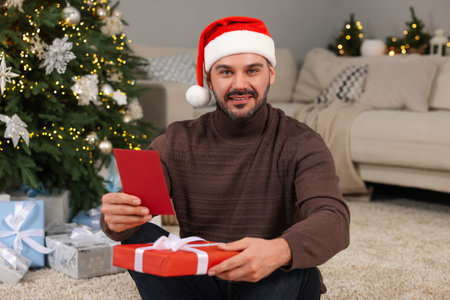Happy man in Santa hat with Christmas gift and greeting card at homeの写真素材