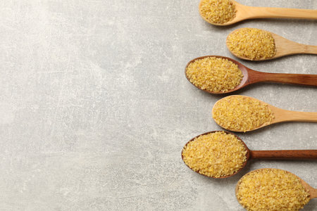 Spoons with raw bulgur on gray table, flat lay. Space for text