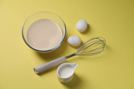 Metal whisk, dough in bowl, eggs and milk on pale yellow background, above view
