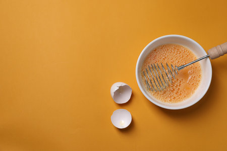 Beaten eggs, metal whiskey in bowl and shells on orange background, flat lay. Space for textの写真素材