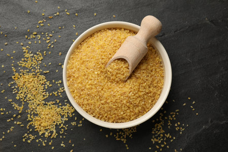 Bowl and scoop with raw bulgur on dark gray table, top view