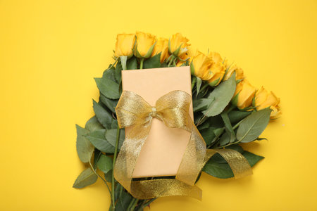 Gift box with golden bow and bouquet of beautiful roses on yellow background, flat layの写真素材