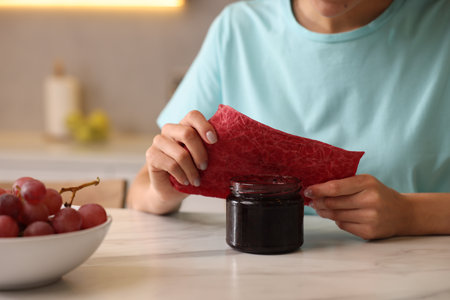 Woman packing jar of jam into beeswax food wrap at light table in kitchen, closeupの写真素材