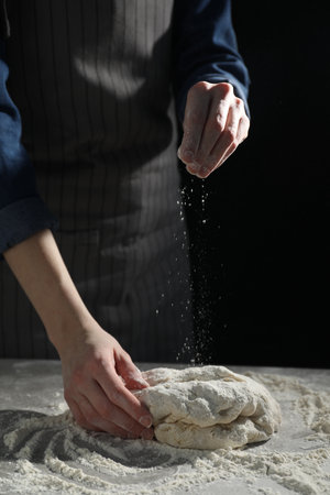 Making bread. Woman sprinkling flour over dough at table on dark background, closeupの写真素材