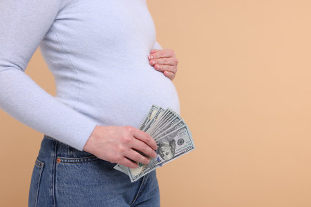 Surrogate mother. Pregnant woman with dollar banknotes on beige background, closeup. Space for textの写真素材