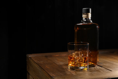 Whiskey with ice cubes in glass and bottle on wooden crate against black background, space for textの写真素材
