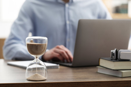 Hourglass with flowing sand on desk. Man using laptop indoors, selective focusの写真素材