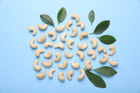 Many tasty cashew nuts and leaves on light blue background, flat lay