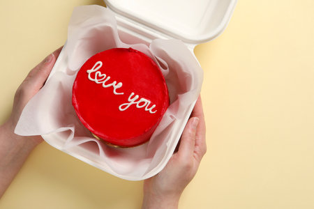 Woman holding takeaway box with bento cake at beige table, top view. St. Valentine's day surprise