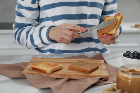 Woman spreading tasty nut butter onto toast at table, closeup