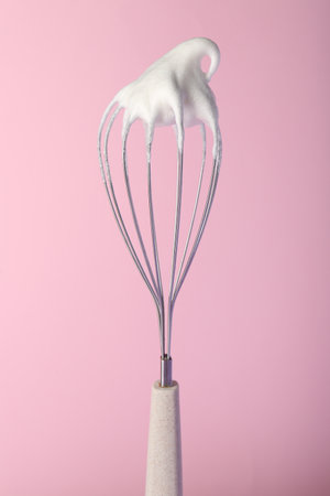 Whisk with whipped egg whites on pink background