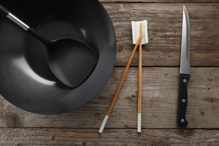 Black metal wok, chopsticks, knife and spatula on wooden table, flat lay