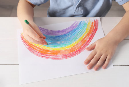 Little boy drawing rainbow with pencil at white wooden table indoors, closeup. Child`s artの写真素材