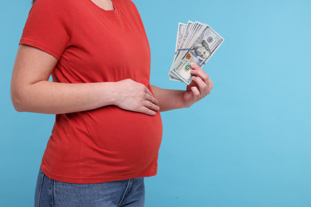Surrogate mother. Pregnant woman with dollar banknotes on light blue background, closeup. Space for textの写真素材