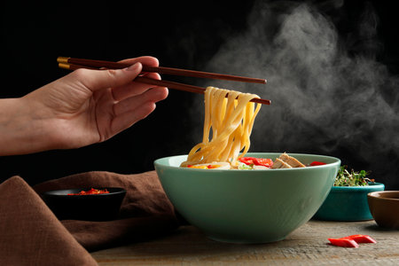 Woman eating delicious ramen with chopsticks at wooden table, closeup. Noodle soupの写真素材