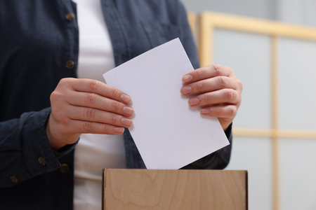 Woman putting her vote into ballot box on blurred background, closeup