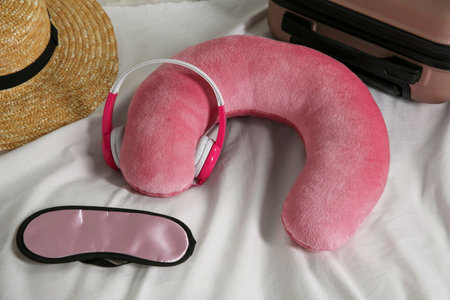 Pink travel pillow with headphones, suitcase, hat and sleep mask on bedの写真素材