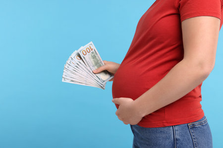 Surrogate mother. Pregnant woman with dollar banknotes on light blue background, closeup. Space for textの写真素材