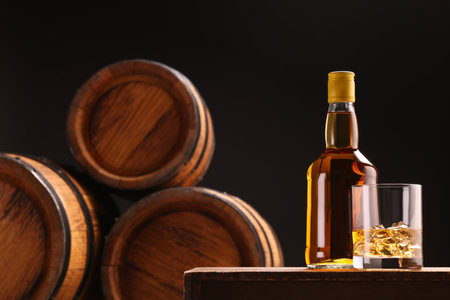 Whiskey with ice cubes in glass and bottle on wooden table near barrels against black background, space for textの写真素材