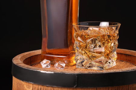 Whiskey in glass, bottle and ice cubes on wooden barrel against black backgroundの写真素材