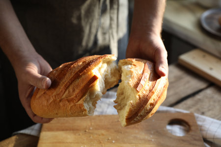 Man breaking loaf of fresh bread at wooden table indoors, closeup