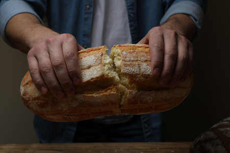 Man breaking loaf of fresh bread at wooden table, closeup
