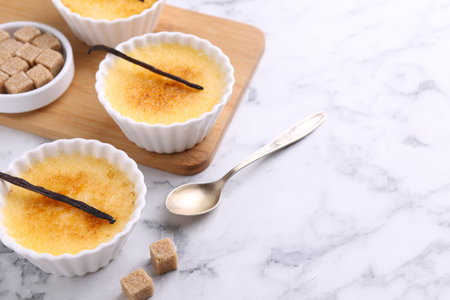 Delicious creme brulee in bowls, vanilla pods, sugar cubes and spoon on white marble table, space for textの写真素材