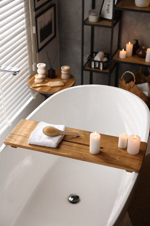 Wooden tray with burning candles, towel and brush on bathtub in bathroomの写真素材
