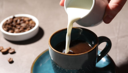 Woman pouring milk into cup with aromatic coffee at light table, closeupの写真素材