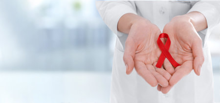 Cancer awareness. Doctor holding red ribbon on blurred background, closeup. Banner design with space for textの写真素材