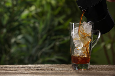 Pouring coffee over ice in glass cup at wooden table outdoors. Space for textの写真素材