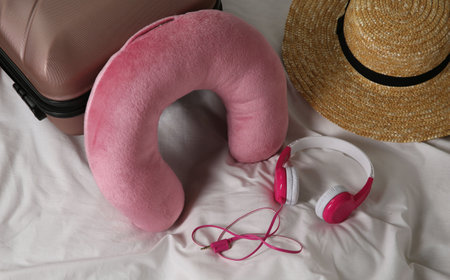 Pink travel pillow, suitcase, hat and headphones on bedの写真素材