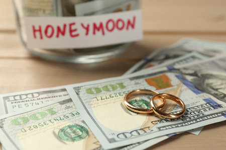 Glass jar with word Honeymoon, dollar banknotes and golden rings on wooden table, closeupの写真素材